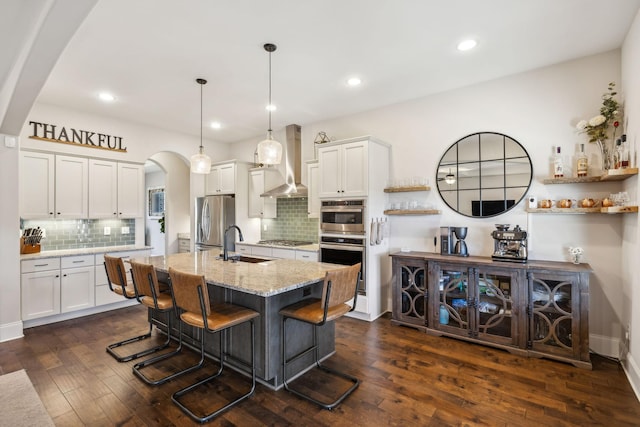 kitchen featuring arched walkways, white cabinets, stainless steel appliances, wall chimney range hood, and a sink