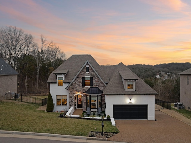 view of front of home with a yard, roof with shingles, fence, stone siding, and concrete driveway