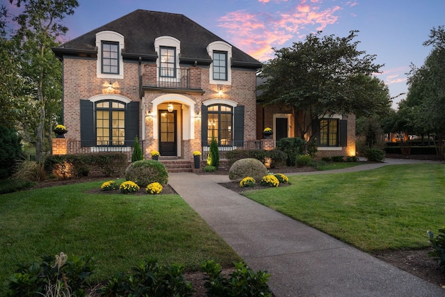 french country home featuring roof with shingles, brick siding, and a lawn