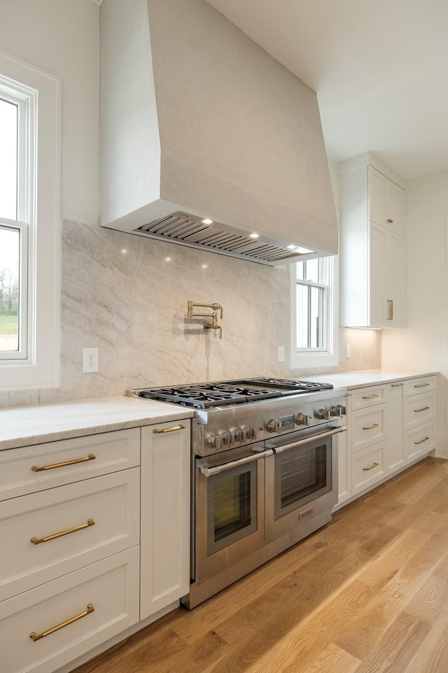 kitchen featuring double oven range, light wood finished floors, tasteful backsplash, premium range hood, and white cabinets
