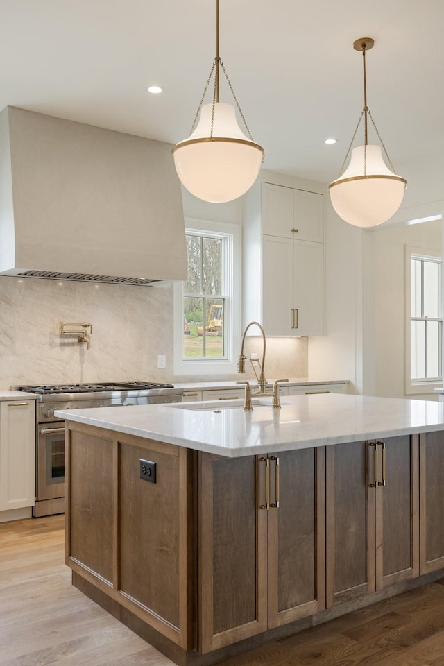 kitchen featuring white cabinetry, light wood-style flooring, premium range hood, range with two ovens, and a kitchen island with sink