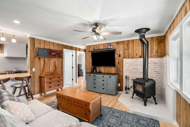 living room with wooden walls, light wood-style flooring, ceiling fan, a wood stove, and crown molding