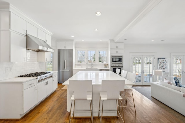 kitchen with under cabinet range hood, stainless steel appliances, wood finished floors, a kitchen breakfast bar, and french doors