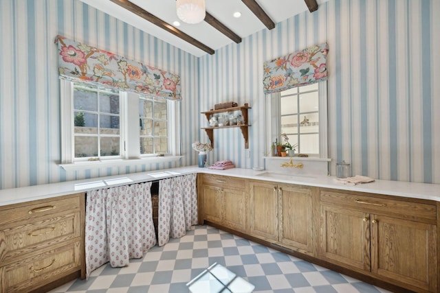 kitchen with brown cabinetry, beamed ceiling, light floors, and wallpapered walls