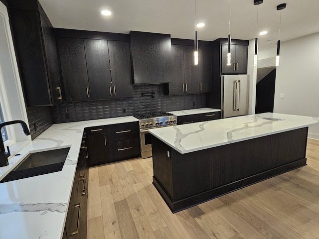 kitchen with high end appliances, light wood-style flooring, a sink, wall chimney range hood, and dark cabinets