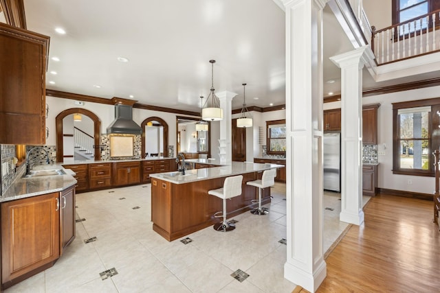 kitchen with stainless steel appliances, a sink, wall chimney range hood, decorative backsplash, and decorative columns