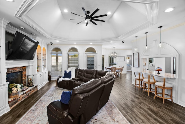 living room featuring dark wood finished floors, a raised ceiling, ornamental molding, a stone fireplace, and ceiling fan with notable chandelier