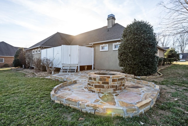 rear view of house featuring an outdoor fire pit, a lawn, a chimney, roof with shingles, and stucco siding