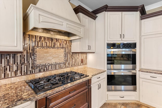 kitchen featuring double oven, black gas stovetop, light stone counters, backsplash, and custom range hood