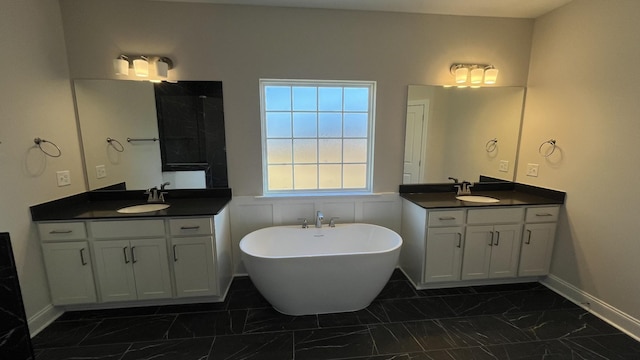 full bath with marble look tiles, a soaking tub, two vanities, and baseboards