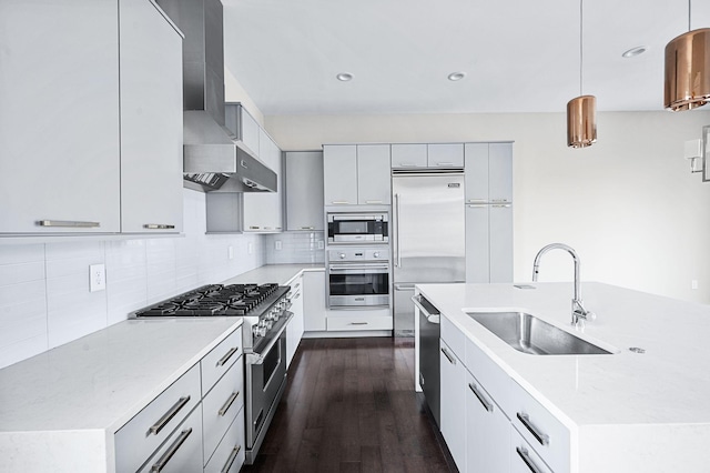 kitchen with pendant lighting, backsplash, a sink, built in appliances, and wall chimney exhaust hood