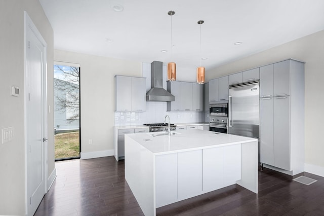 kitchen with dark wood-style floors, tasteful backsplash, gray cabinets, wall chimney range hood, and built in appliances