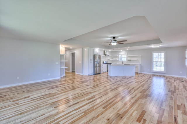 unfurnished living room with a tray ceiling, baseboards, ceiling fan, and light wood finished floors