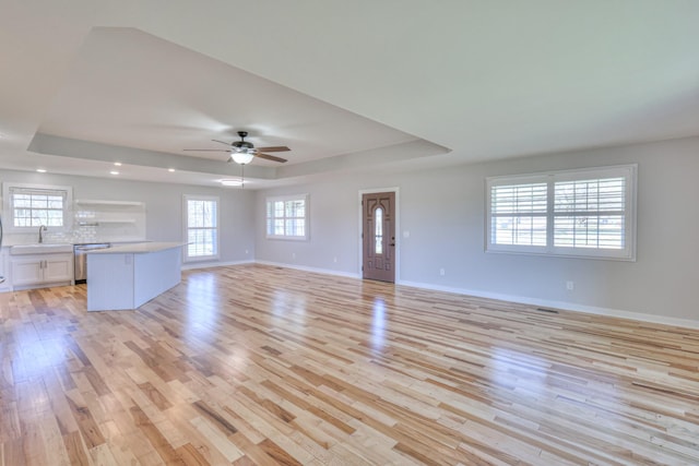 unfurnished living room with a raised ceiling, baseboards, a sink, and light wood finished floors