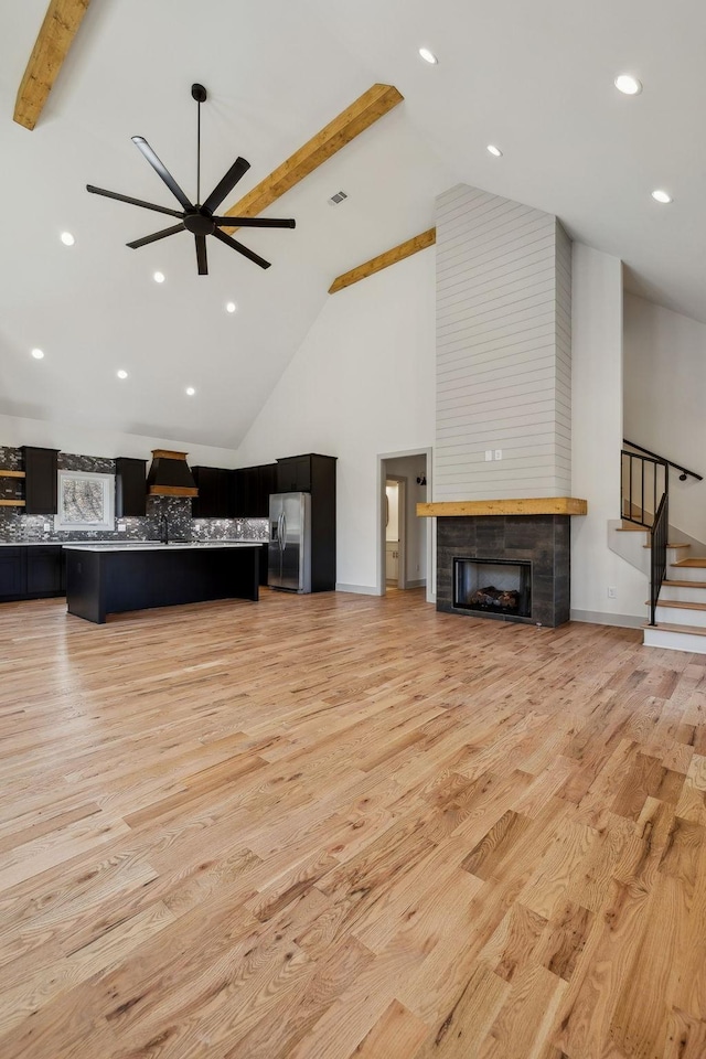 living room featuring a ceiling fan, a tiled fireplace, beamed ceiling, stairs, and light wood-style floors