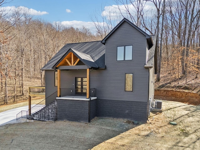 view of front facade featuring a shingled roof, a wooded view, and central air condition unit