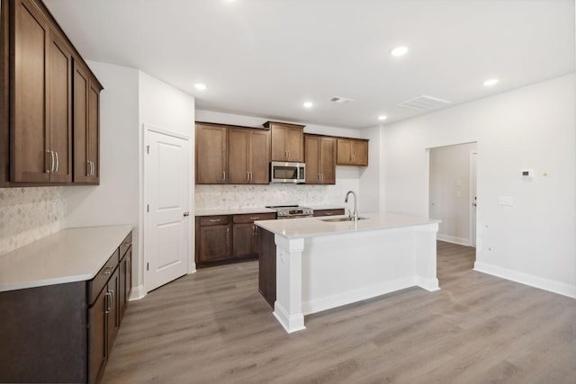 kitchen featuring a center island with sink, a sink, stainless steel appliances, light wood-type flooring, and backsplash