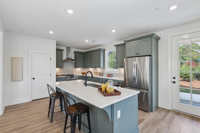 kitchen featuring stainless steel appliances, a sink, wall chimney range hood, backsplash, and light wood finished floors