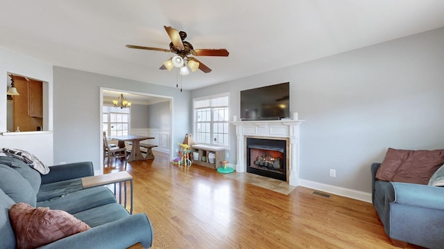 living area featuring visible vents, light wood-style flooring, a fireplace with flush hearth, baseboards, and ceiling fan with notable chandelier