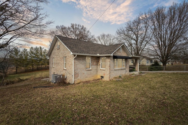 property exterior at dusk featuring cooling unit, brick siding, fence, a yard, and roof with shingles