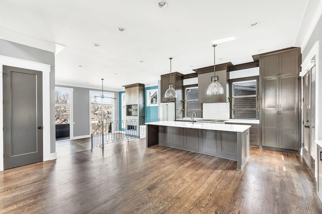 kitchen with dark wood-style floors, pendant lighting, light countertops, decorative backsplash, and a kitchen island with sink