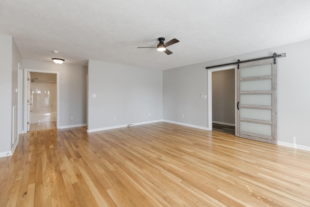 unfurnished room with a barn door, a ceiling fan, a textured ceiling, light wood-type flooring, and baseboards