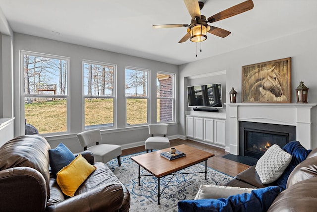 living area featuring a fireplace with flush hearth, baseboards, ceiling fan, and dark wood-type flooring