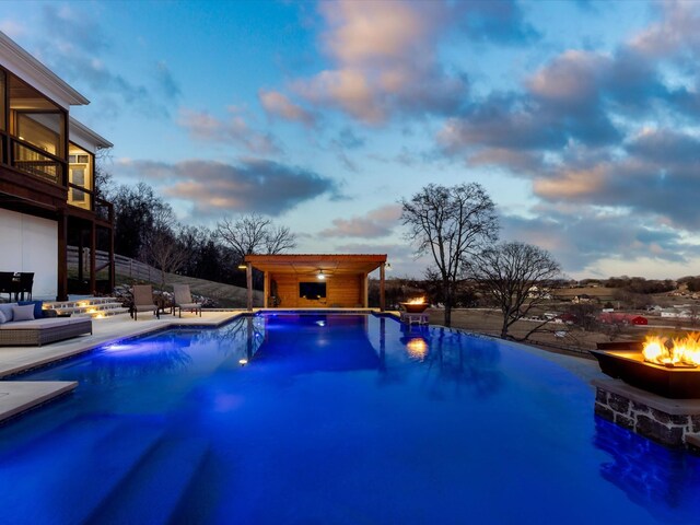 pool at dusk with ceiling fan, a patio, an outdoor living space with a fire pit, and an outdoor pool