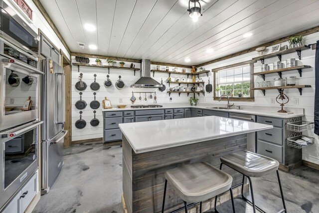 kitchen with wall chimney exhaust hood, stainless steel appliances, gray cabinetry, open shelves, and a sink