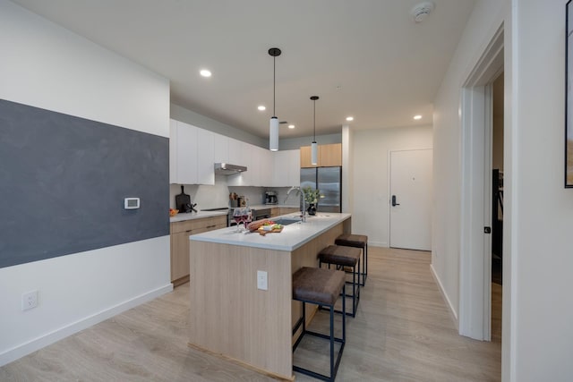 kitchen with a breakfast bar, light countertops, a kitchen island with sink, under cabinet range hood, and stainless steel refrigerator