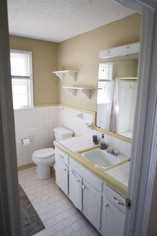 full bath with a textured ceiling, toilet, vanity, tile walls, and wainscoting