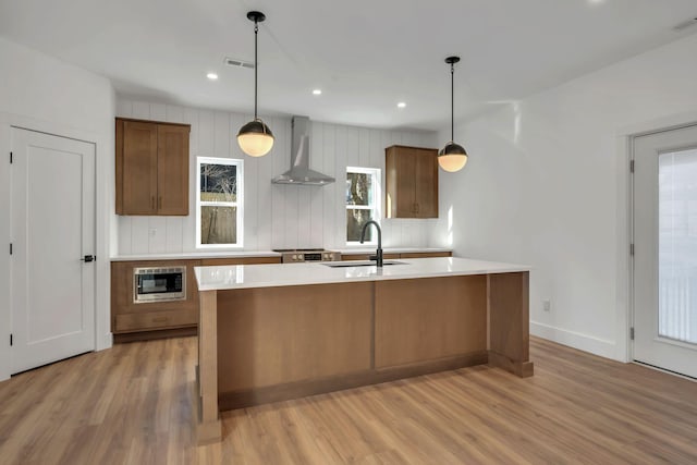 kitchen featuring light wood finished floors, an island with sink, light countertops, wall chimney range hood, and a sink