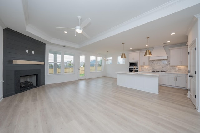 unfurnished living room with light wood-style flooring, a large fireplace, a ceiling fan, ornamental molding, and a raised ceiling