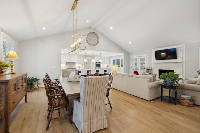 dining area with light wood finished floors, beamed ceiling, a brick fireplace, high vaulted ceiling, and a notable chandelier