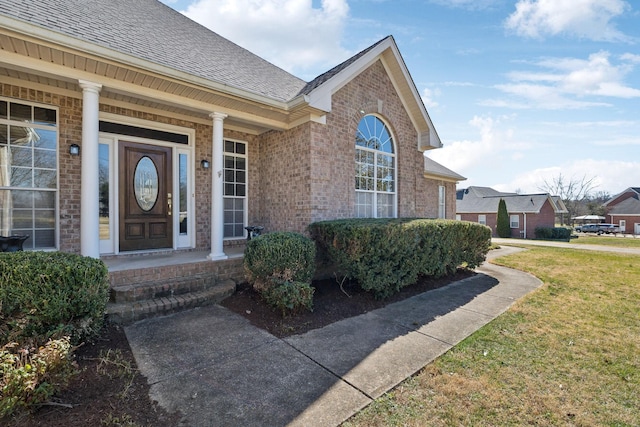 view of exterior entry with a yard, brick siding, and roof with shingles