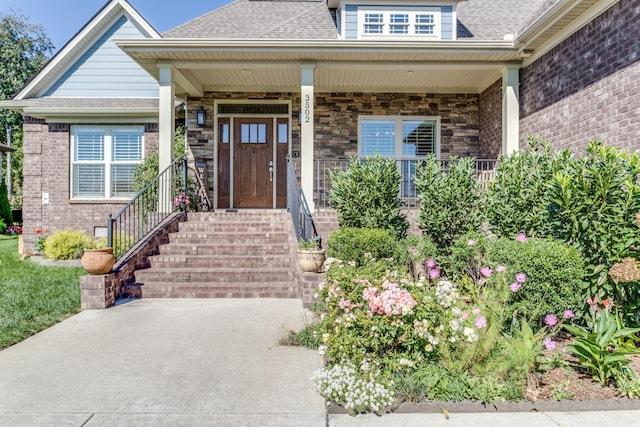 view of exterior entry featuring covered porch, stone siding, a shingled roof, and brick siding