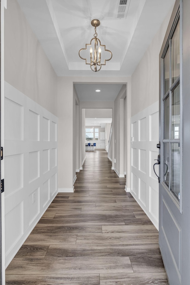 hall featuring a chandelier, a decorative wall, wood finished floors, visible vents, and a raised ceiling