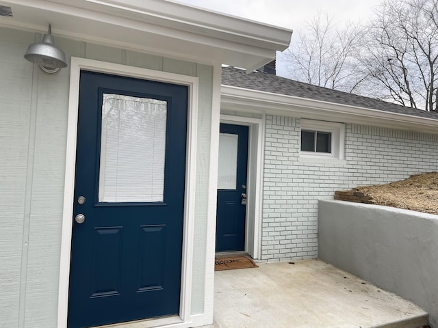 view of exterior entry featuring roof with shingles, brick siding, and visible vents