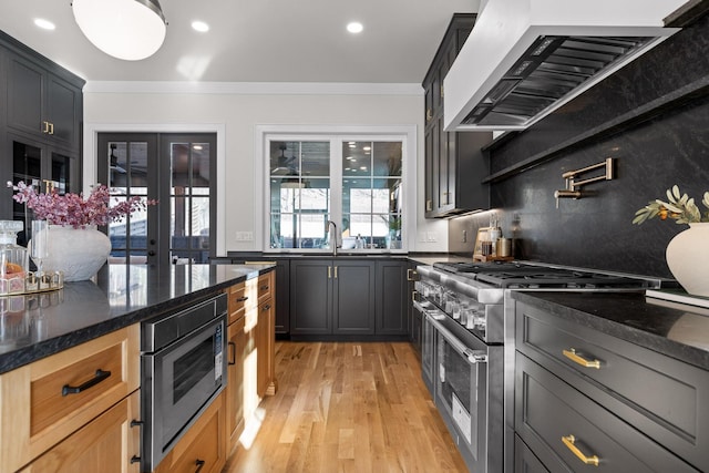 kitchen with stainless steel appliances, ornamental molding, light wood-type flooring, range hood, and dark stone countertops