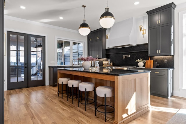 kitchen with french doors, a breakfast bar area, dark countertops, light wood-style flooring, and premium range hood