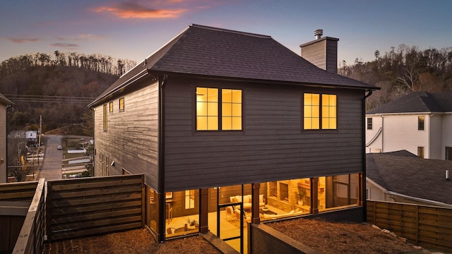 back of property at dusk featuring a shingled roof, a chimney, and fence