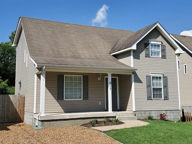 traditional-style home with covered porch, a front yard, fence, and roof with shingles