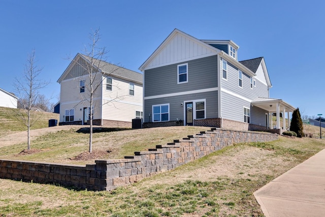 view of front facade with board and batten siding, brick siding, a front lawn, and central AC unit