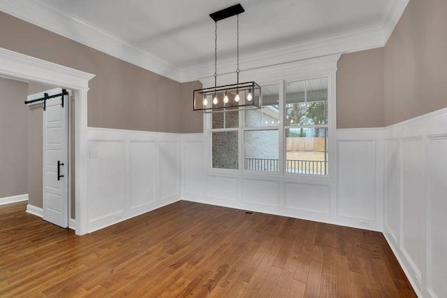 unfurnished dining area featuring a barn door, wainscoting, wood finished floors, crown molding, and a decorative wall