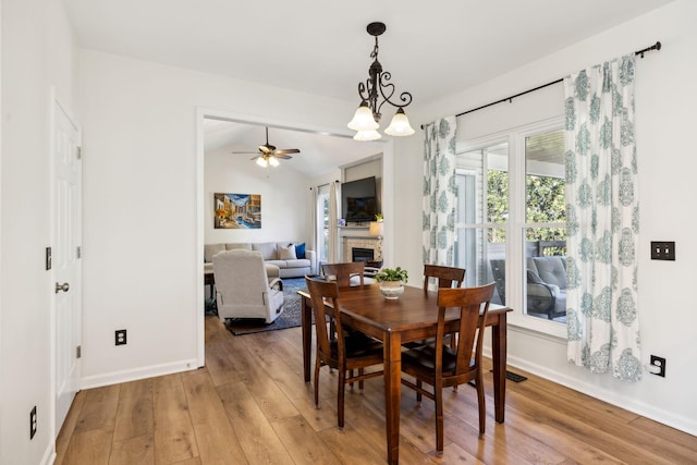 dining area with light wood-style flooring, a glass covered fireplace, vaulted ceiling, baseboards, and ceiling fan with notable chandelier