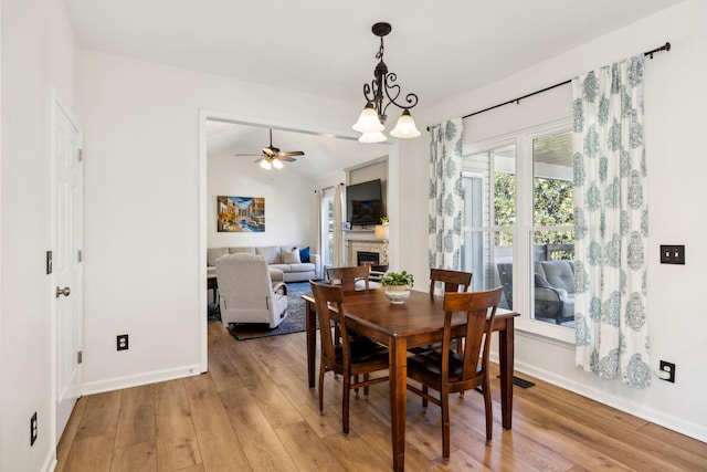 dining space with light wood finished floors, baseboards, vaulted ceiling, a fireplace, and ceiling fan with notable chandelier