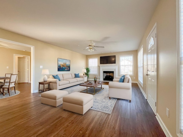 living area with a ceiling fan, a fireplace with flush hearth, baseboards, and dark wood-style flooring