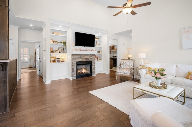 living room with ceiling fan, a stone fireplace, built in shelves, dark wood-type flooring, and crown molding