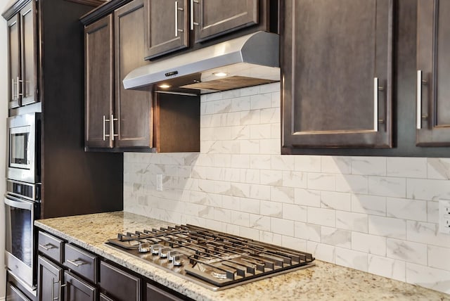 kitchen with light stone counters, stainless steel appliances, dark brown cabinets, under cabinet range hood, and backsplash