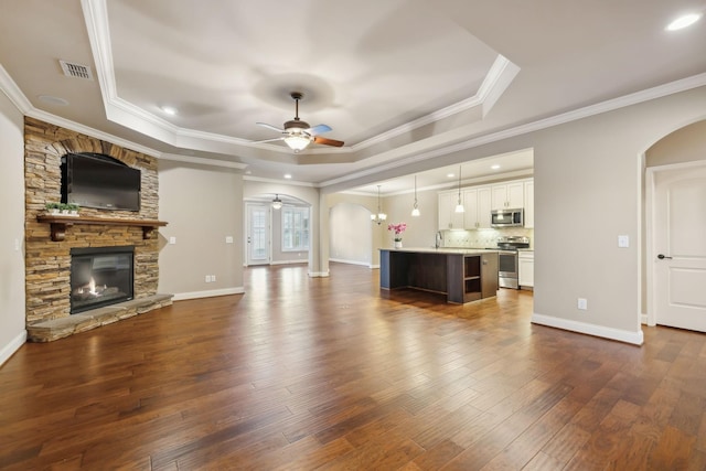 unfurnished living room featuring arched walkways, a raised ceiling, ceiling fan, dark wood-type flooring, and a stone fireplace